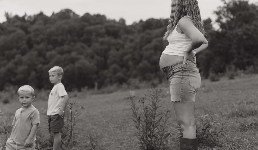 An expecting mother stands in a field on her property admiring her older two boys playing during a maternity session in Boone North Carolina.