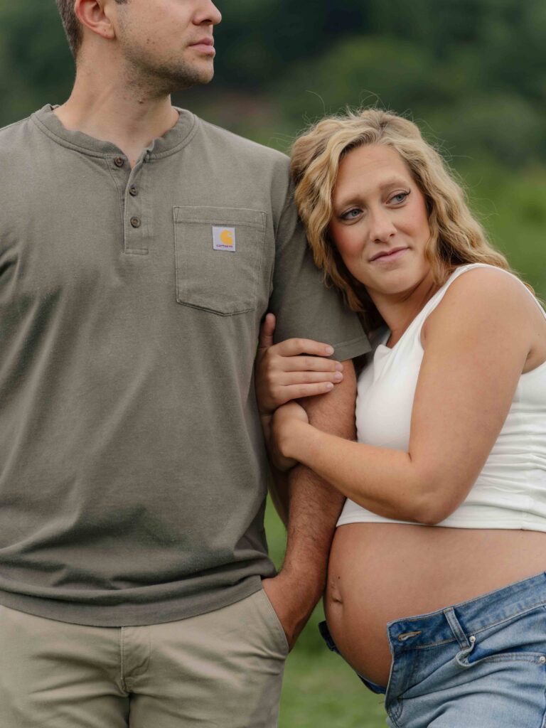 Mom and dad stand in an open field on their family property in Boone North Carolina during their maternity session, looking off at their two boys playing with their cows in the pasture. 