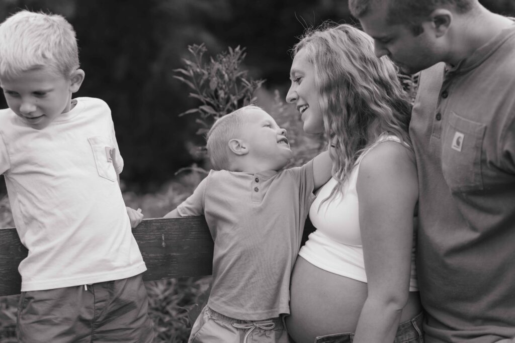 Little boy puts a flower in mom's hair in between poses during a lifestyle family maternity photoshoot in Boone North Carolina while dad and older brother lean on a fence.