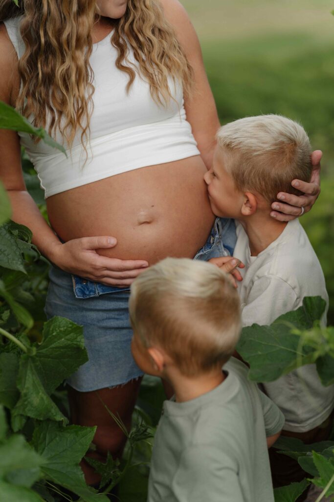 Oldest brother kisses his mom's belly during a lifestyle family maternity session in Boone North Carolina in the garden of their property.