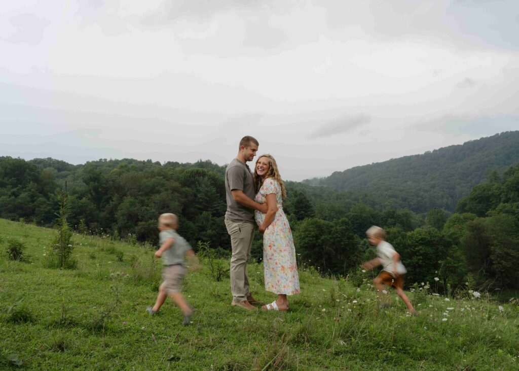 Mom and dad stand together and laugh for a directed photo in the field of their family property as their two boys run circles around them during a maternity session in Boone North Carolina.