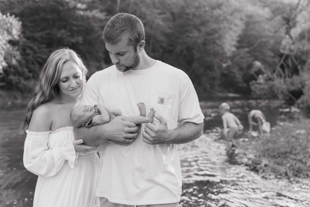 Mom and dad admire their newborn son together as dad cradles the baby in the middle of the river in Boone North Carolina while their older two sons play in the river.