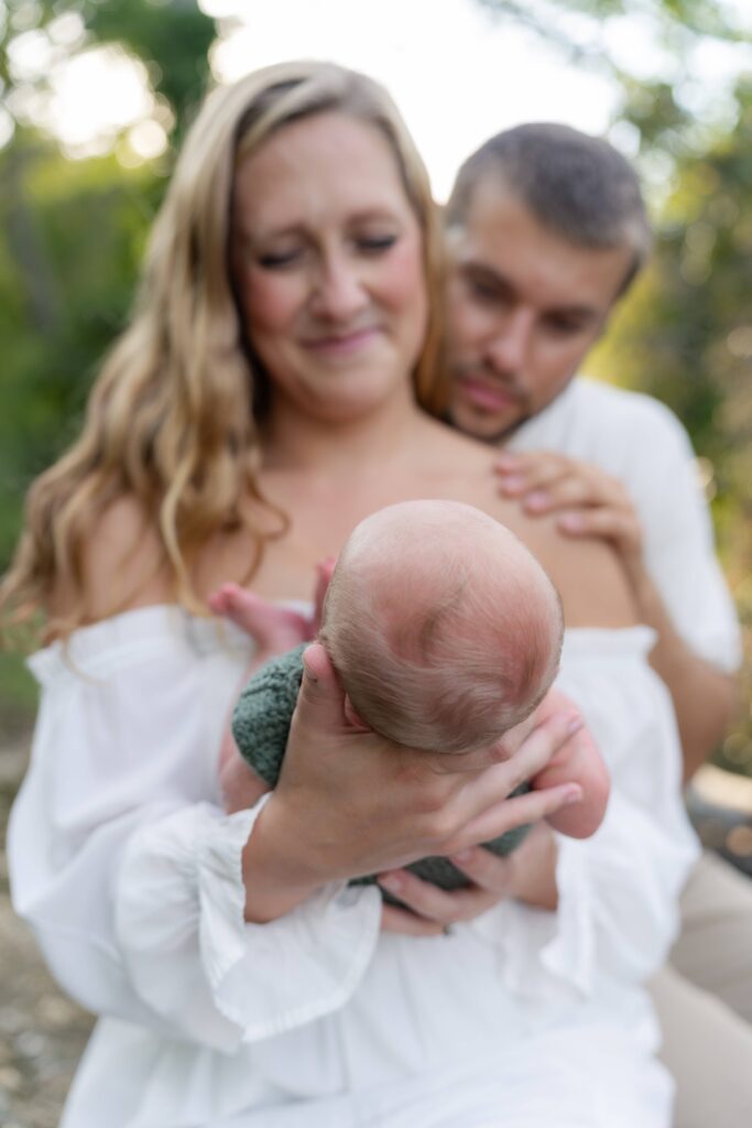 Mom and dad admire their 3rd son during a lifestyle newborn photoshoot while sitting on a hand-made bench in the river of their Boone NC property.