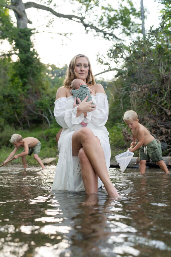 Mother holds her newborn son while sitting on a hand-made bench in the river along their family property in Boone NC while her two older boys play in the river.
