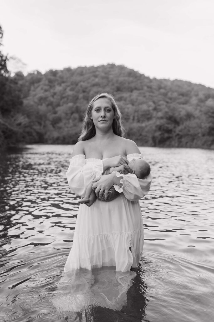New mom of 3 boys stands in the middle of a river on her property in Boone North Carolina cradling her newborn son during a lifestyle newborn photoshoot.